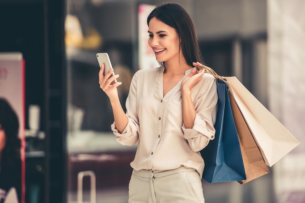 Uma mulher carregando sacolas de compras sorrindo e olhando para o celular.