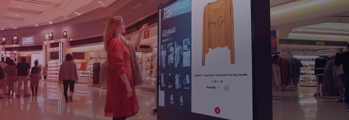 Uma mulher de vestido vermelho olha para uma tela digital que mostra um moletom amarelo. O moletom tem a escrita "Hey Girl" impressa nele. A tela exibe o moletom e os tamanhos disponíveis para compra…