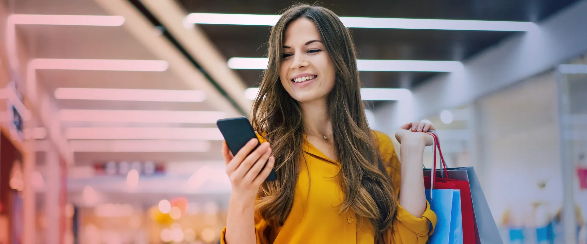 A imagem mostra uma jovem sorridente segurando um smartphone na mão esquerda e várias sacolas de compras na mão direita. Ela está vestida com uma camisa amarela e tem cabelos longos e castanhos. O fu…