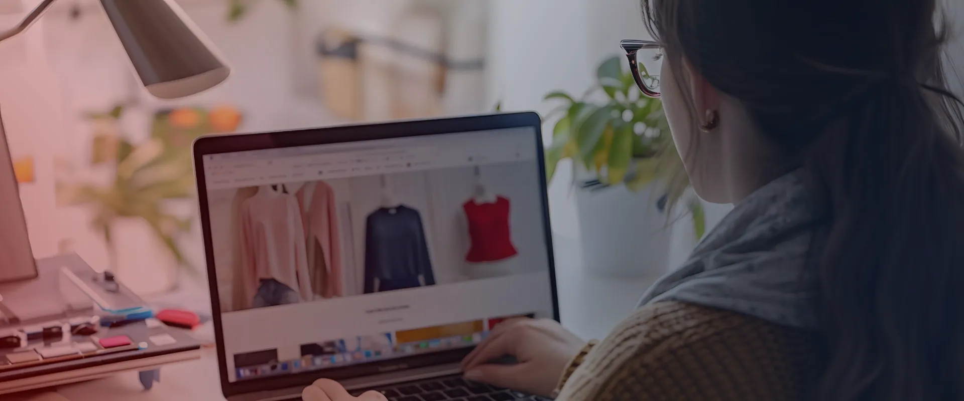 Uma mulher está sentada à frente de um laptop e olha para a tela. Seu cabelo está escuro e ela está vestindo óculos e uma jaqueta bege. Ela está usando a mão direita para clicar no touchpad do laptop…