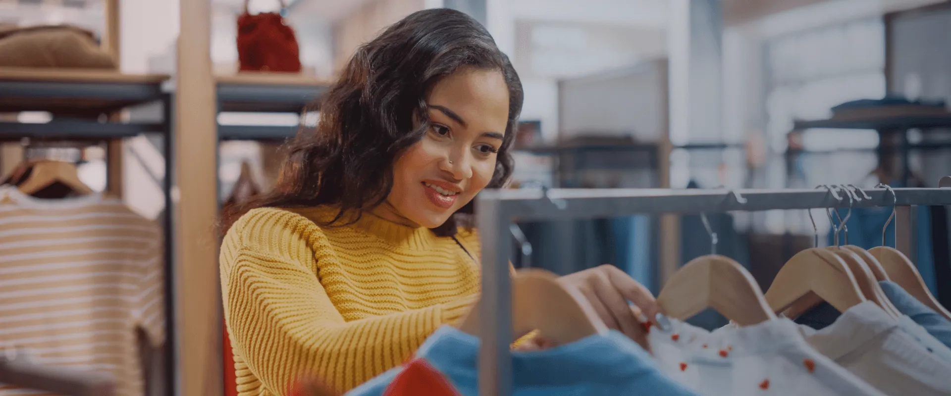 A imagem mostra uma mulher jovem com cabelo escuro e ondulado, vestindo um suéter amarelo, olhando para roupas penduradas em um cabide em uma loja de roupas. Ela está sorrindo e parece estar escolhen…