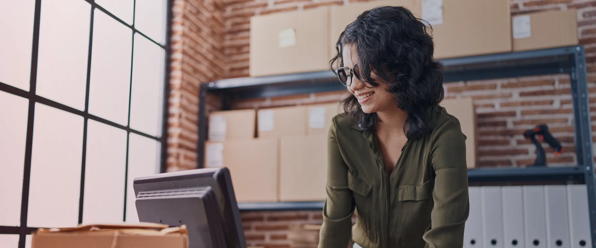 A imagem mostra uma mulher de pé em frente a um computador na frente de uma parede de tijolos. Ela está usando óculos e uma camisa verde. Ela está sorrindo para o computador, enquanto parece estar tr…