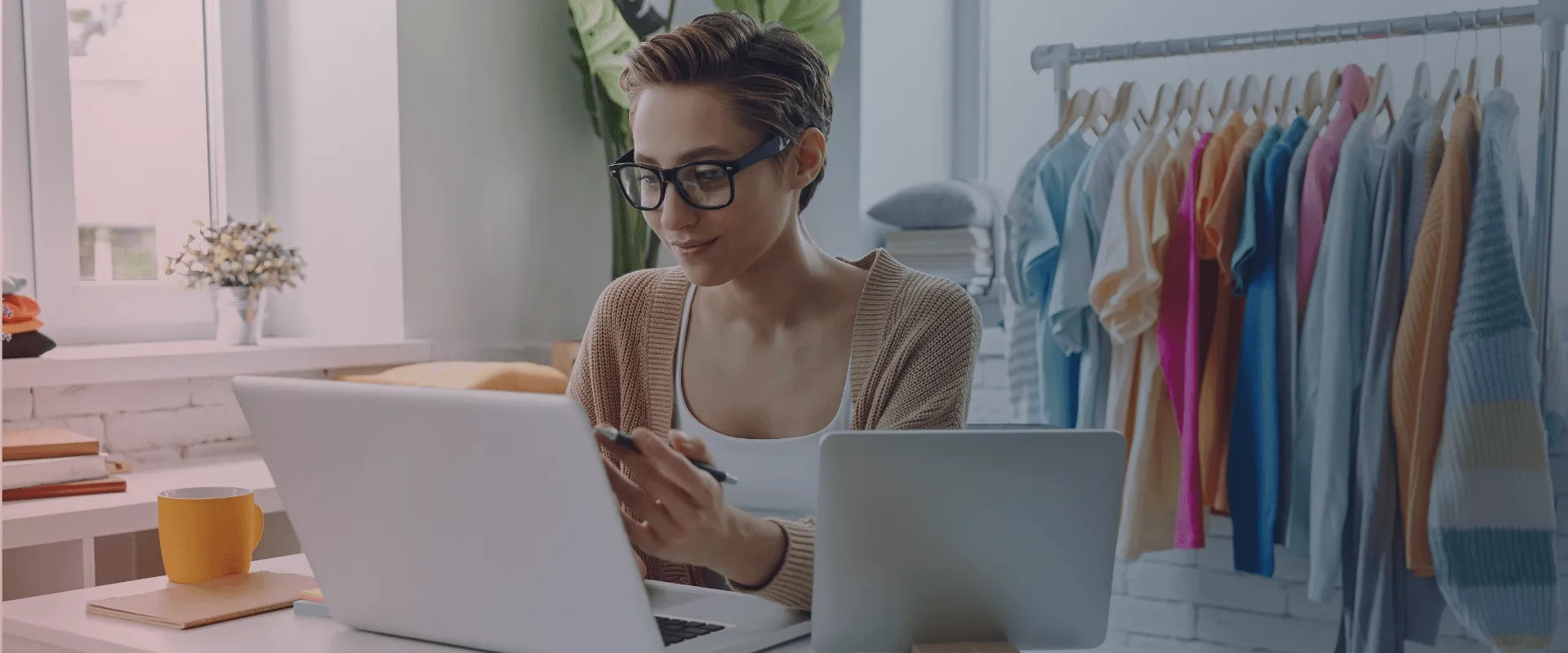 A imagem mostra uma mulher jovem, usando óculos e um suéter bege, sentada à mesa em frente a um laptop. Ela está olhando para o laptop e segurando uma caneta. Há uma xícara amarela e um bloco de nota…