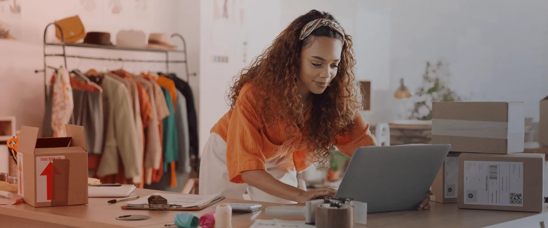 A imagem mostra uma mulher com cabelo encaracolado e uma faixa de cabeça, usando uma blusa laranja e calças brancas, inclinando-se sobre um laptop na mesa. Há uma pilha de caixas de papelão atrás del…