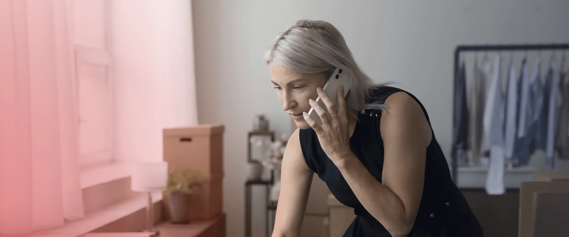 A imagem mostra uma mulher de meia-idade com cabelos grisalhos e curtos, usando um vestido preto, falando ao telefone e digitando em um laptop. Há caixas de papelão e uma arara de roupas atrás dela.…