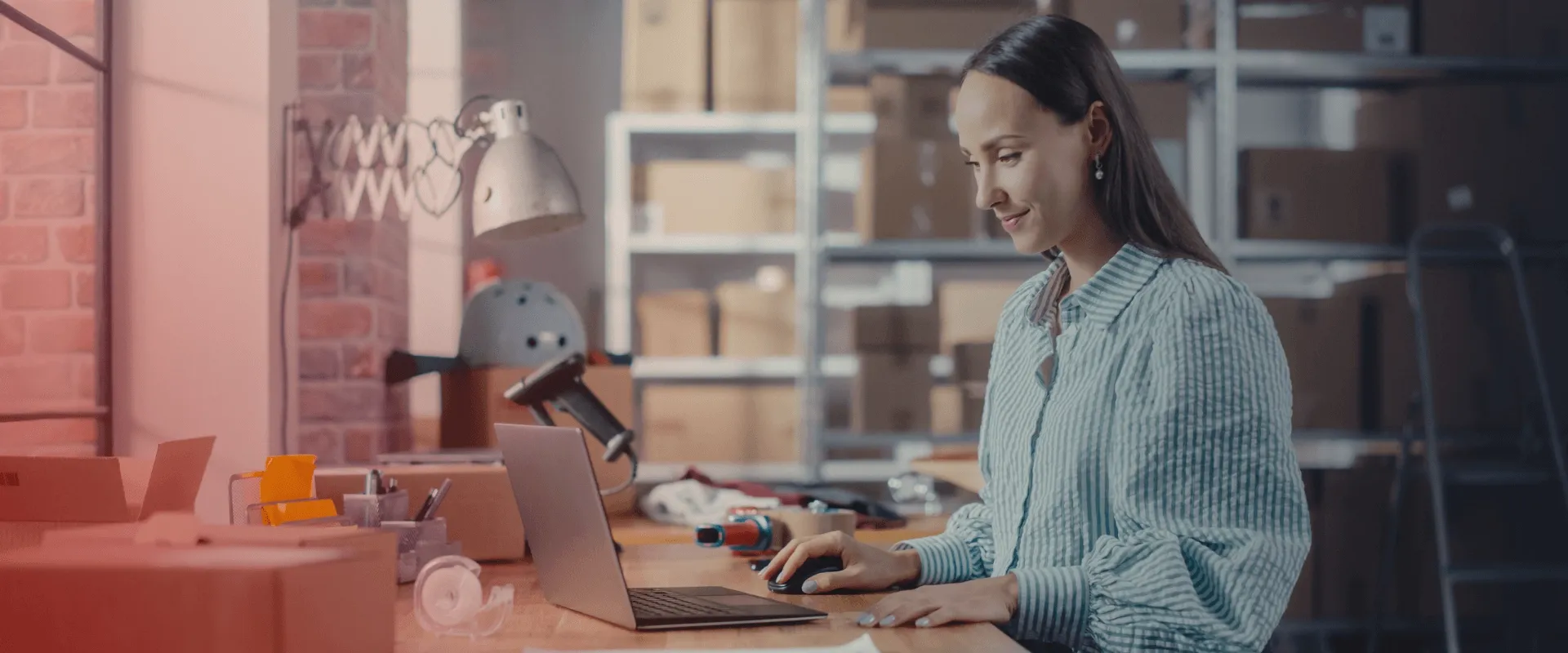 A imagem mostra uma jovem usando uma camisa azul e branca listrada, sentada em frente a um laptop na mesa. Ela está usando um mouse de computador e parece estar trabalhando. Há caixas de papelão atrá…