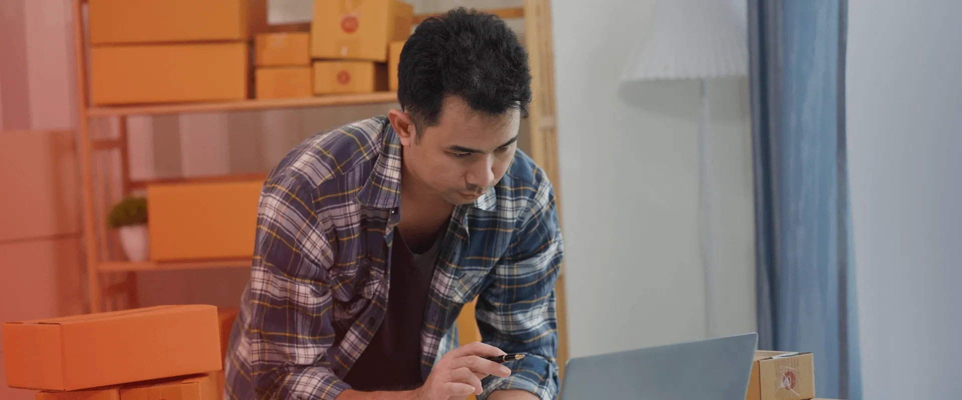 Um jovem está usando um computador na frente de uma prateleira cheia de caixas. Ele está vestindo uma camisa xadrez azul e está se concentrando no computador. Ele parece estar trabalhando em um peque…