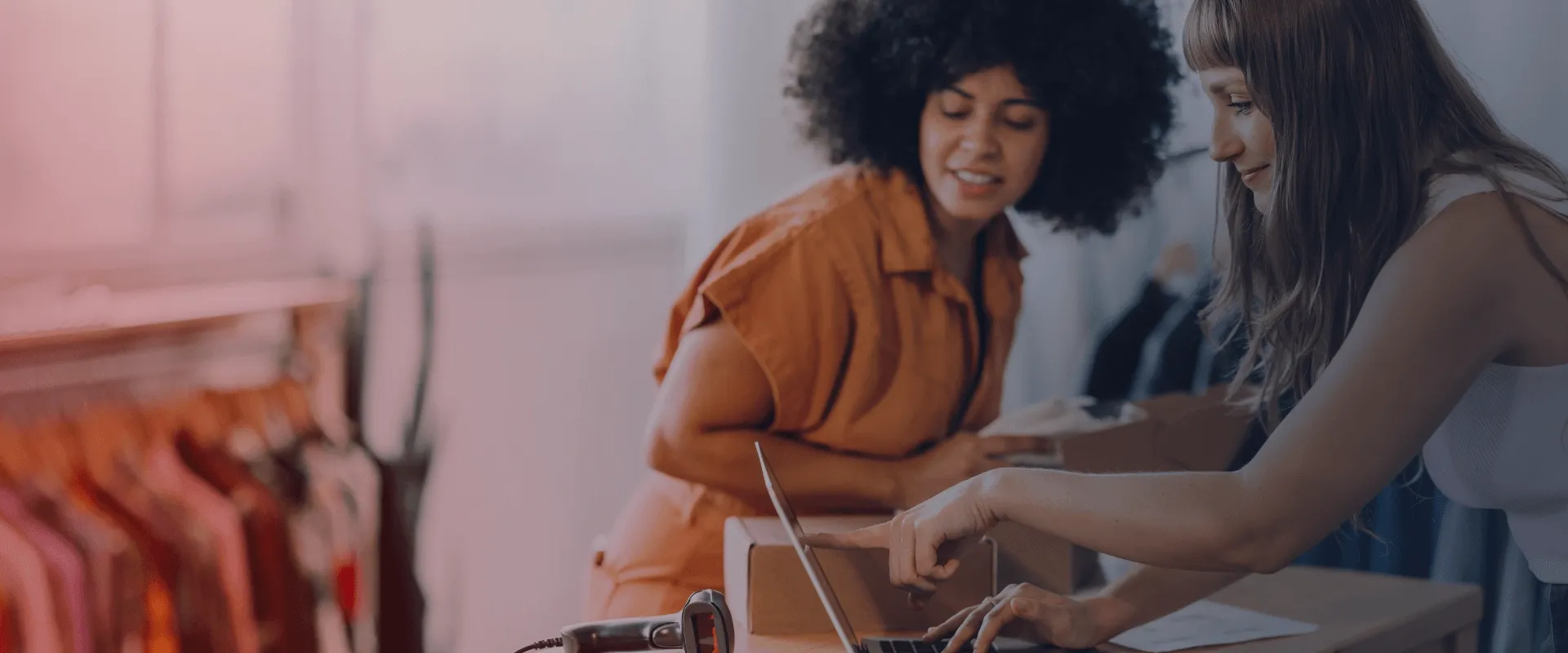 Duas mulheres trabalham juntas em um computador portátil, em um ambiente de loja de roupas.