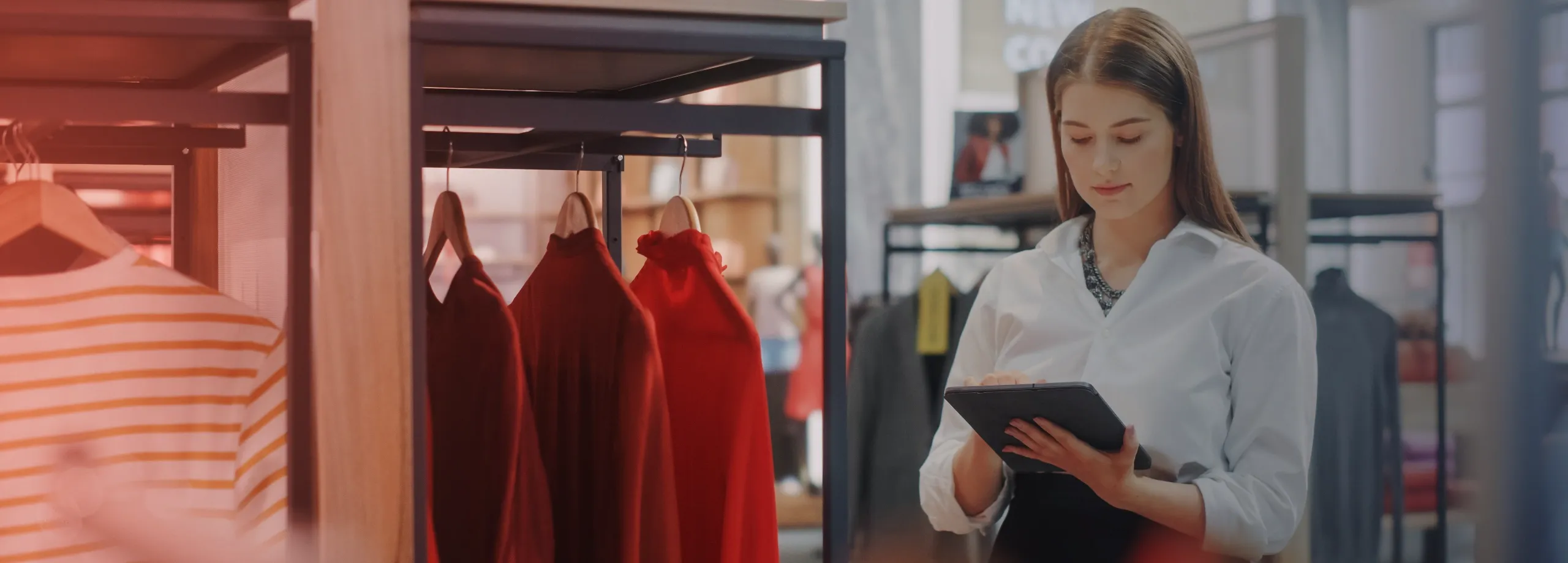 Uma jovem mulher com uma camisa branca está em pé em uma loja de roupas, usando um tablet. Roupas vermelhas e uma camisa listrada estão penduradas em um cabideiro ao fundo.