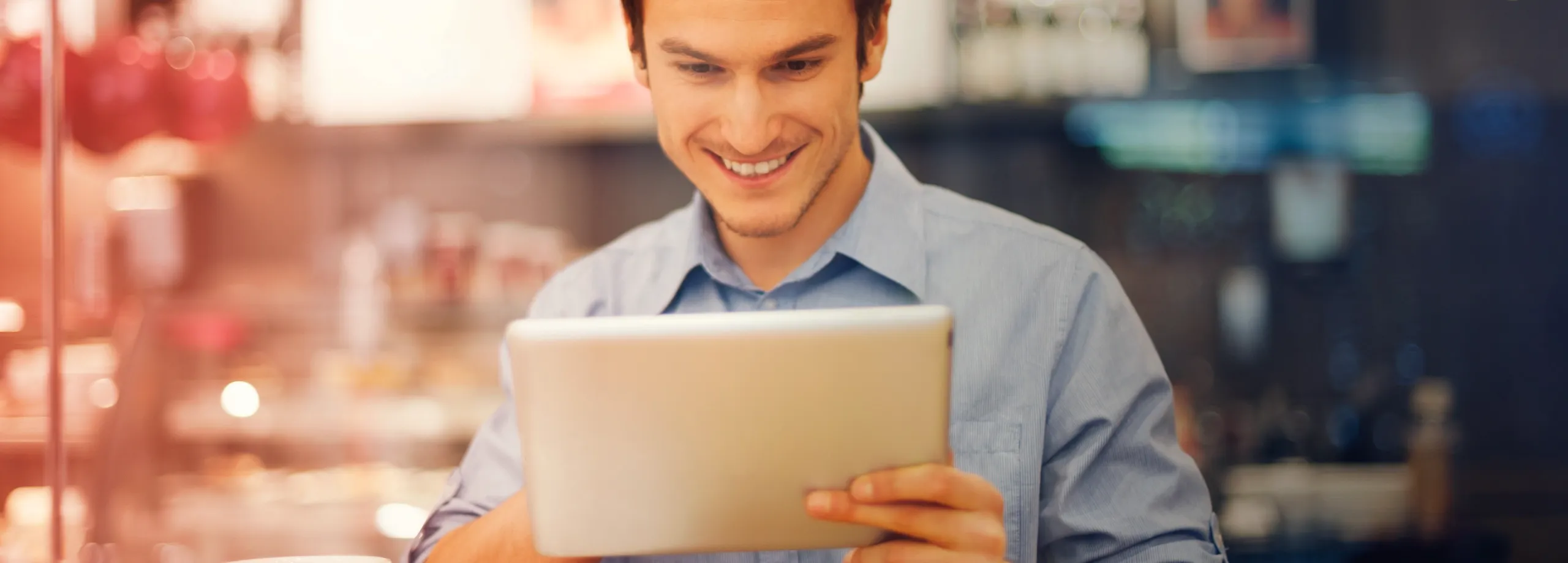 Um homem jovem e sorridente a olhar para um tablet, na frente de um fundo desfocado, que parece ser uma cafetaria ou restaurante.