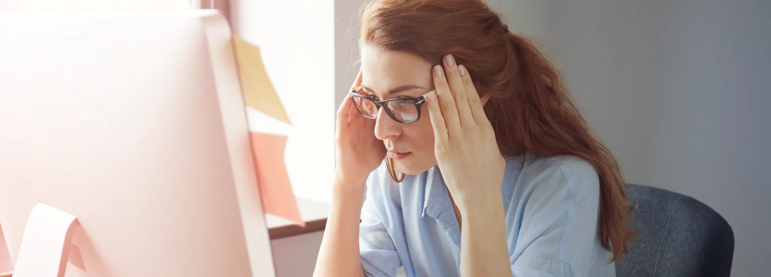 Uma jovem mulher, com a mão na cabeça como se estivesse a ter uma dor de cabeça ou stress, a trabalhar ao computador.