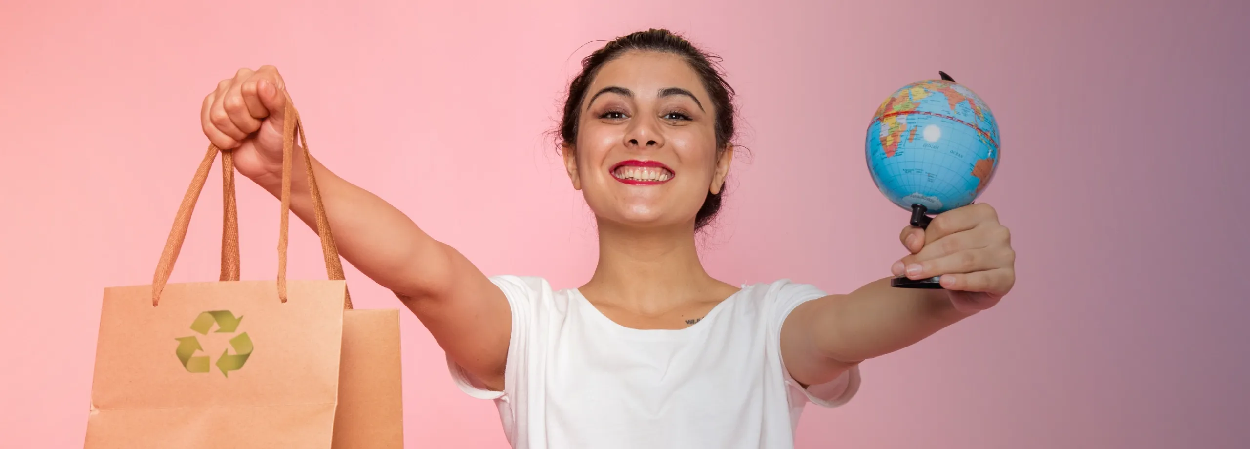 Uma mulher sorrindo, segurando uma sacola de papel com um símbolo de reciclagem em uma mão e em outra um globo terrestre. O fundo é rosa.