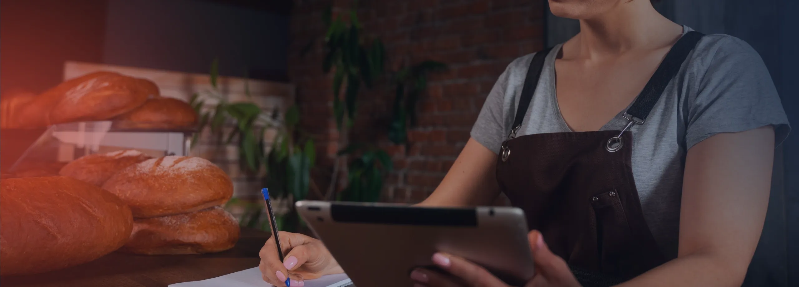 Mulher com camiseta cinza e avental marrom, trabalhando em uma padaria. Ela está sentada atrás de um balcão de madeira, segurando um tablet com uma mão e escrevendo em um caderno com a outra. Vários…