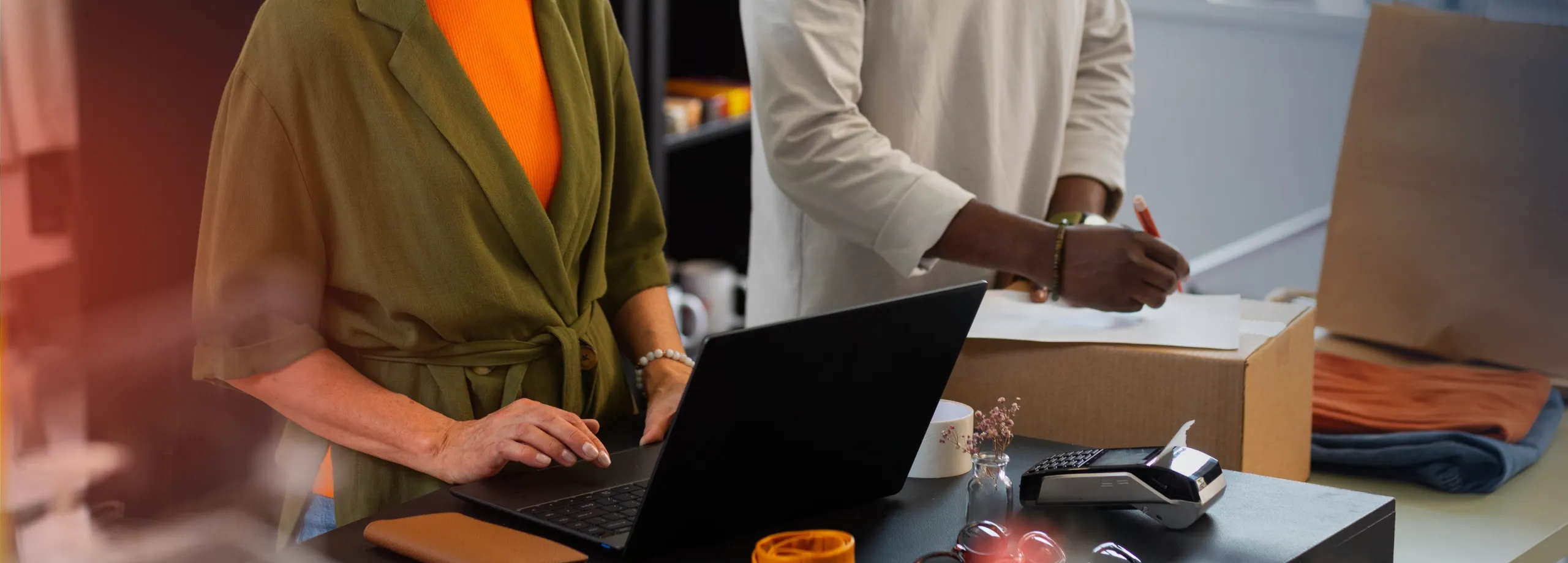 Mulher sorridente, de cabelo curto e escuro, vestindo blusa laranja e casaco verde, usa um laptop em um balcão de loja. Ao lado dela, um homem de blusa clara escreve em um papel sobre uma caixa de pa…