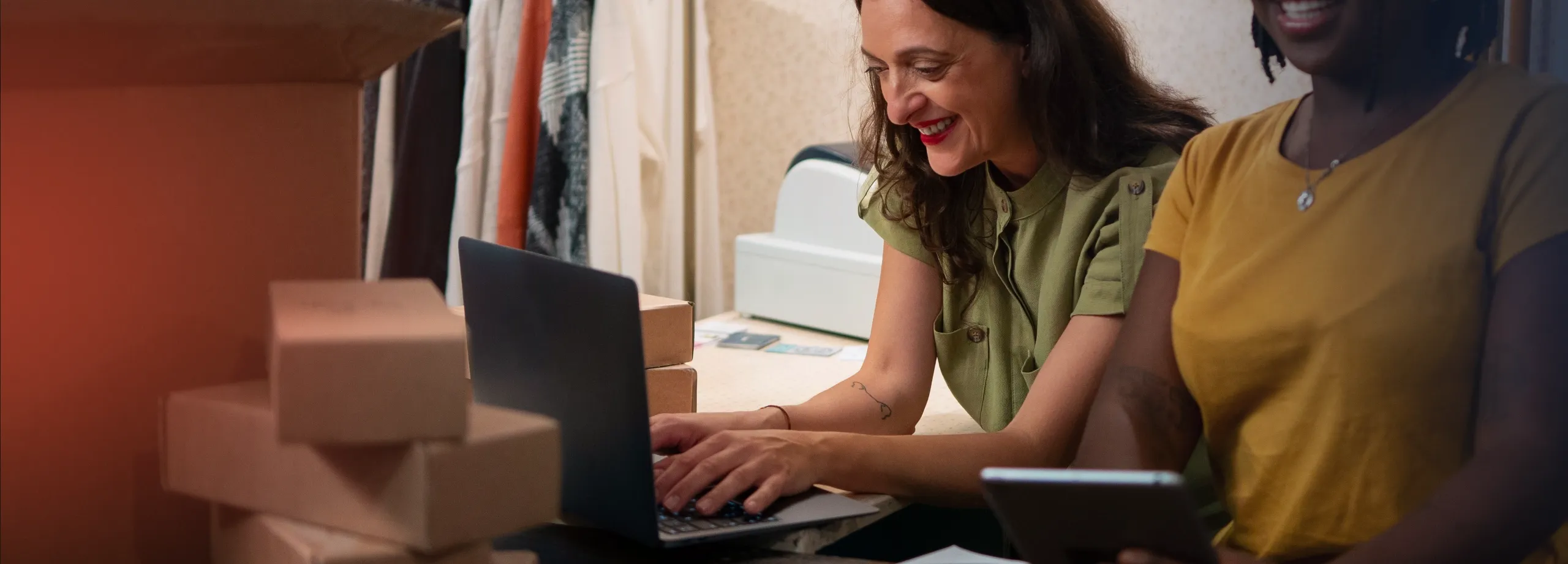 Duas mulheres sorridentes trabalhando juntas em um balcão, provavelmente em uma loja ou expedição de e-commerce. Uma mulher branca, de cabelo escuro e blusa verde, usa um laptop. Ao seu lado, uma mul…