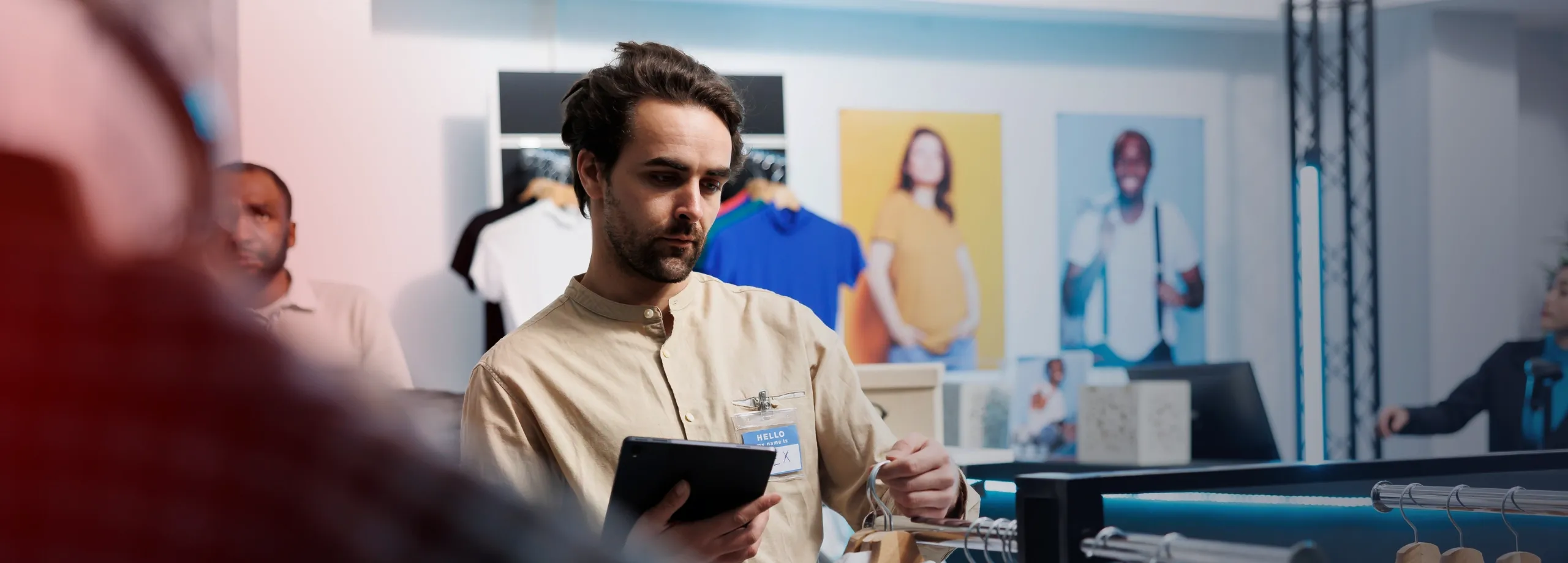 Homem branco, com cabelo escuro e barba, vestindo camisa bege clara e crachá, está em uma loja de roupas. Ele segura um tablet com uma mão enquanto, com a outra, inspeciona uma camisa branca em uma a…