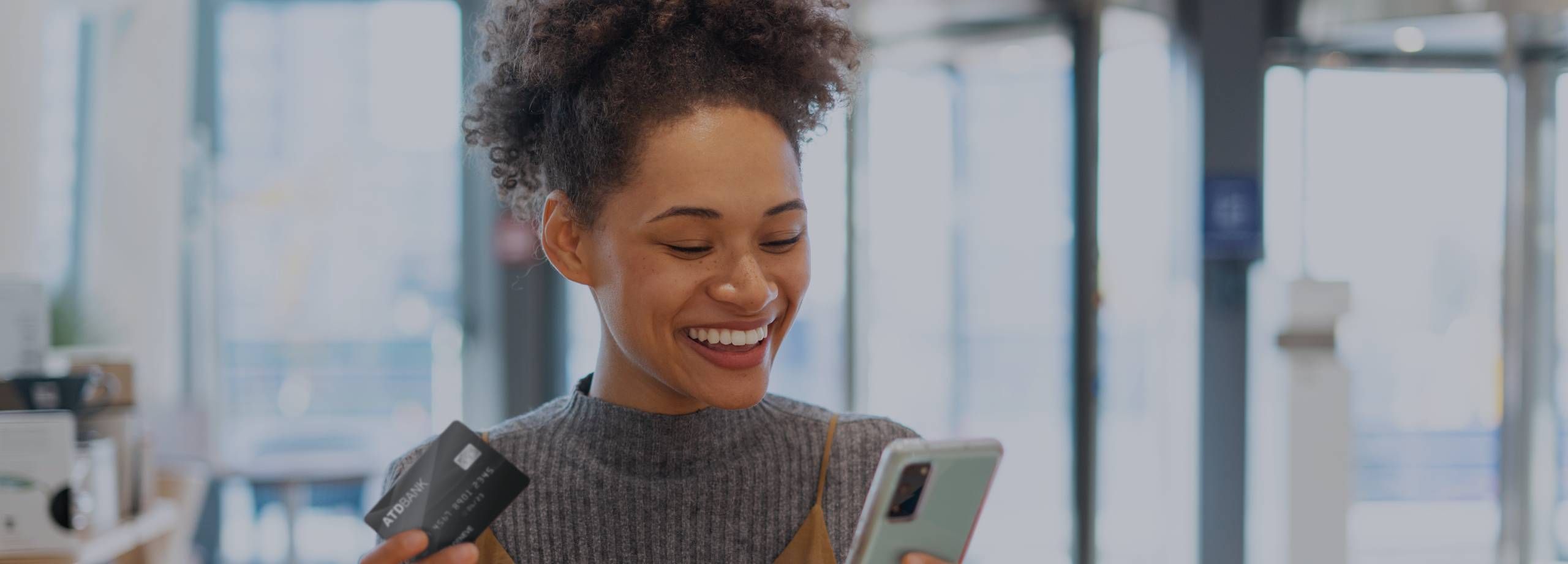 Mulher negra sorridente, com cabelo cacheado preso, vestindo um suéter cinza texturizado sobre uma blusa bege. Ela segura um cartão de crédito preto (ATOBANK) em uma mão e um smartphone na outra, olh…