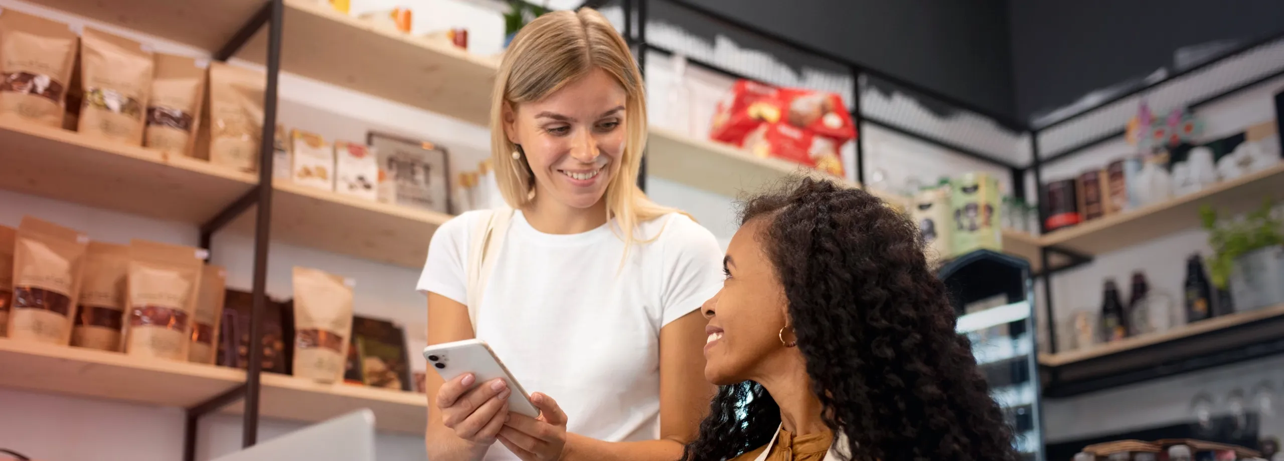 Duas mulheres interagindo em um estabelecimento comercial, possivelmente uma mercearia ou loja de produtos naturais. Uma mulher loira, de camiseta branca e jeans, sorri e mostra a tela de um smartpho…
