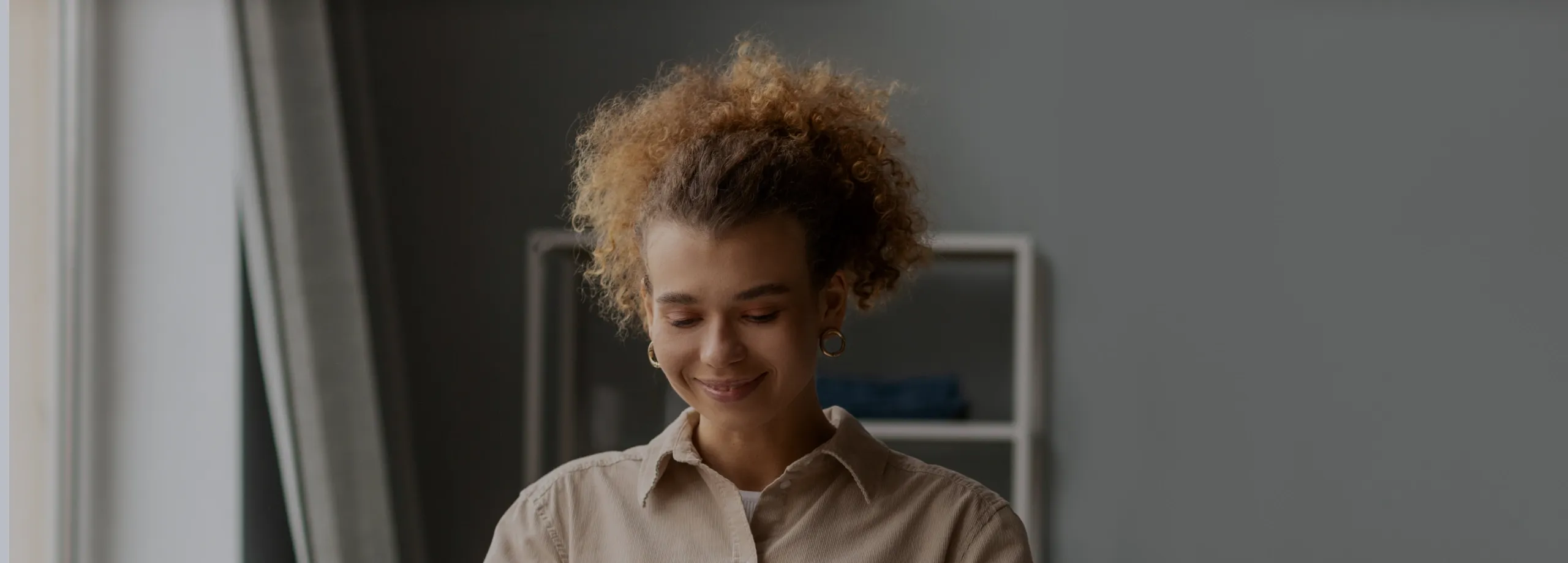 Mulher jovem, de cabelo cacheado claro e volumoso, vestindo camisa de botões bege e calças da mesma cor, em pé em um ambiente que parece ser um estúdio caseiro ou área de expedição de e-commerce. Ela…