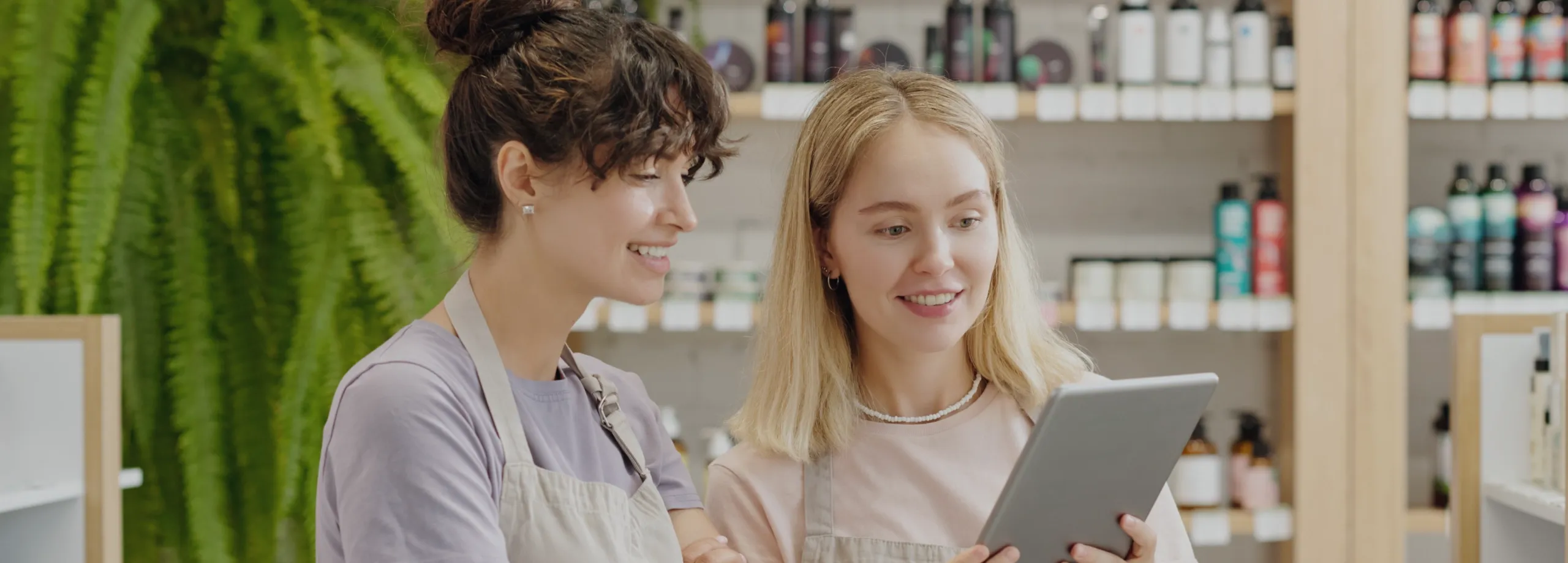 Duas vendedoras sorridentes, ambas de avental, em uma loja de cosméticos ou produtos naturais. Uma mulher loira segura um tablet e mostra o conteúdo para a colega, que tem cabelo escuro preso em um c…