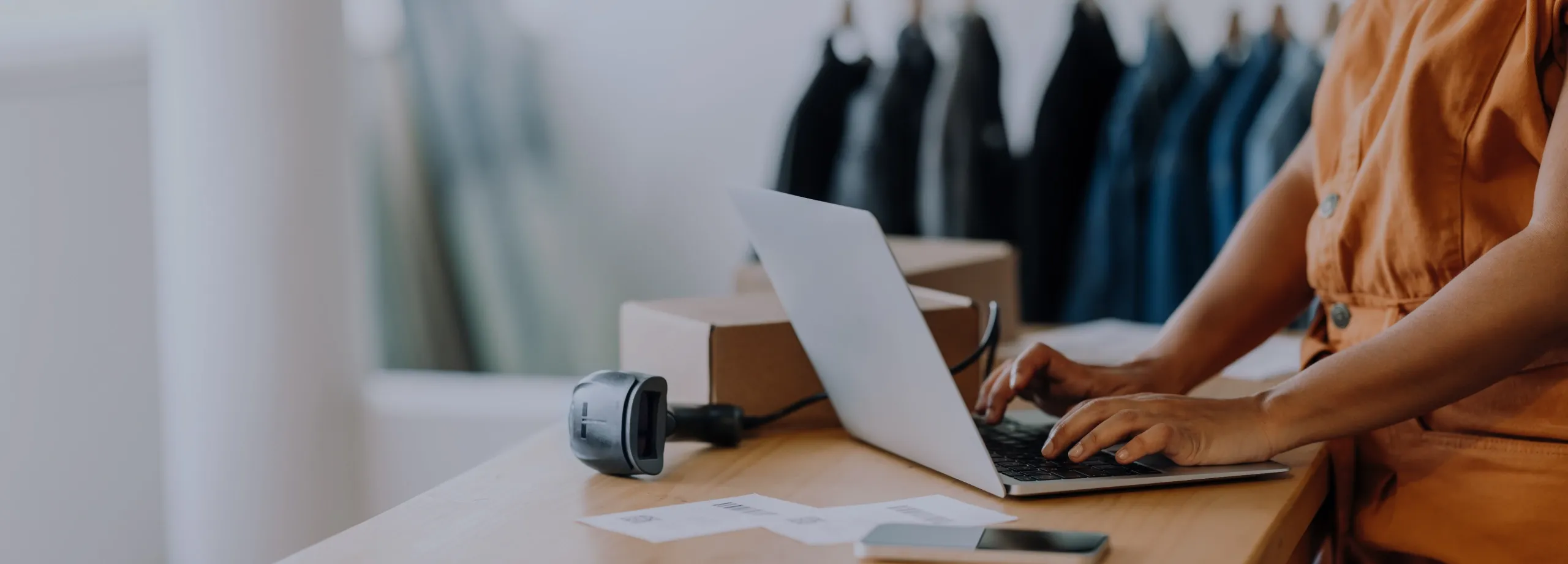 Uma pessoa, possivelmente a dona de um pequeno negócio, está em pé em um balcão de madeira e utiliza um laptop. Na mesa, há também um leitor de código de barras, um smartphone, uma caixa de papelão e…