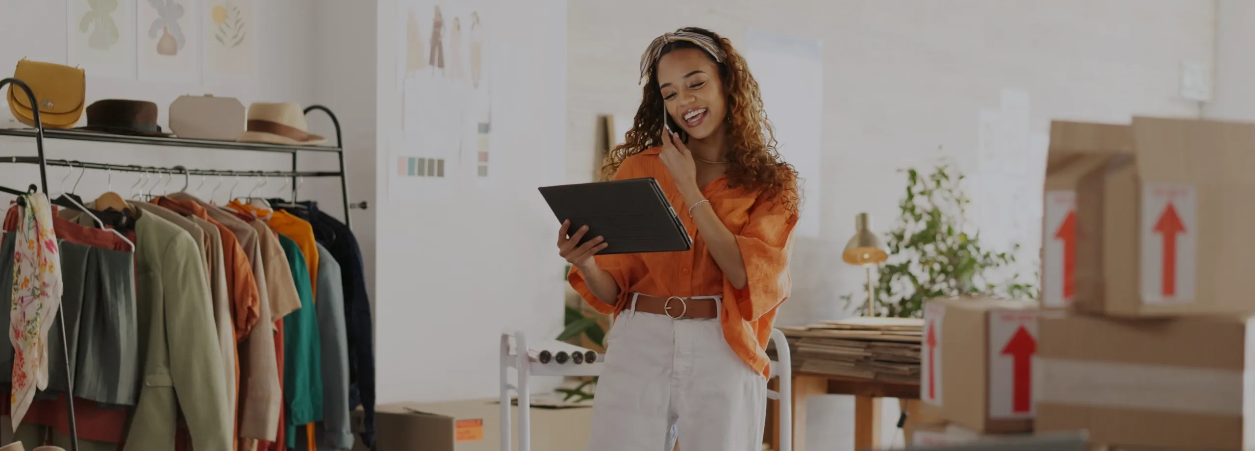 Uma jovem empreendedora de moda, sorrindo, enquanto fala ao celular e olha para um tablet que segura na outra mão. Ela está em pé em seu escritório ou ateliê, vestindo uma camisa laranja e calça bran…