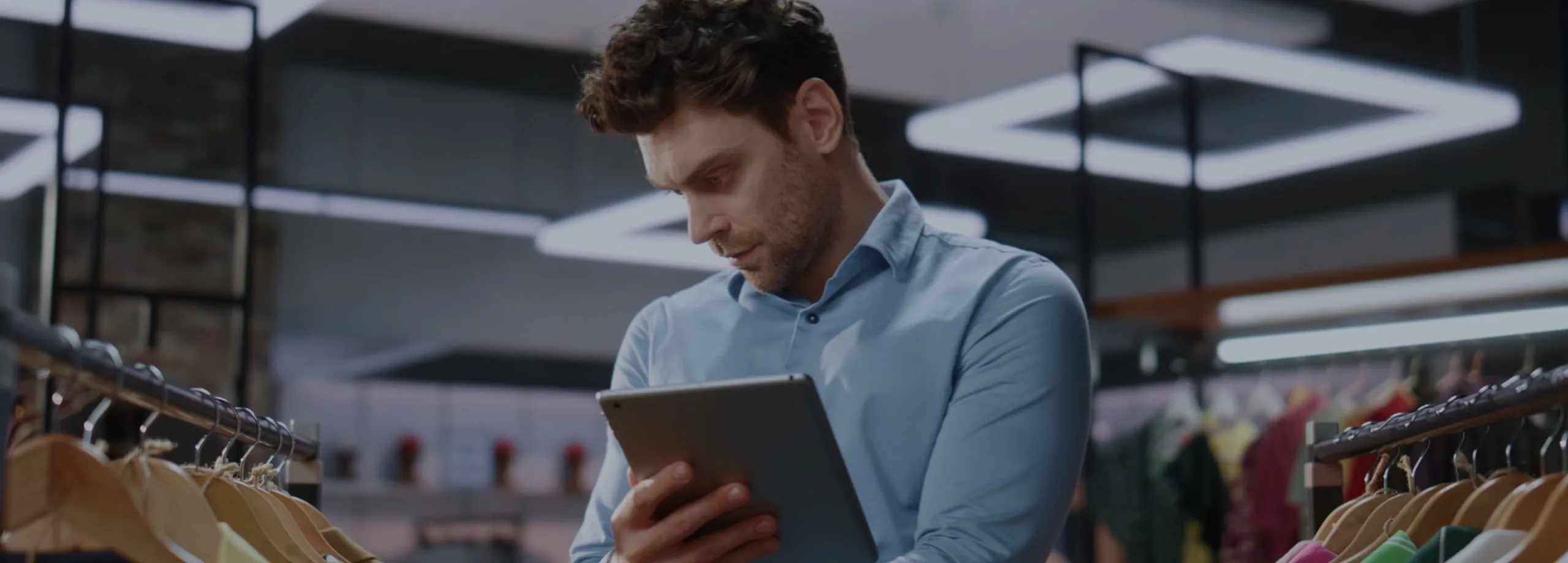 Um homem de camisa azul, possivelmente gerente ou funcionário de uma loja, está em pé em meio a araras de roupas. Ele olha com concentração para um tablet que segura em suas mãos, enquanto verifica o…