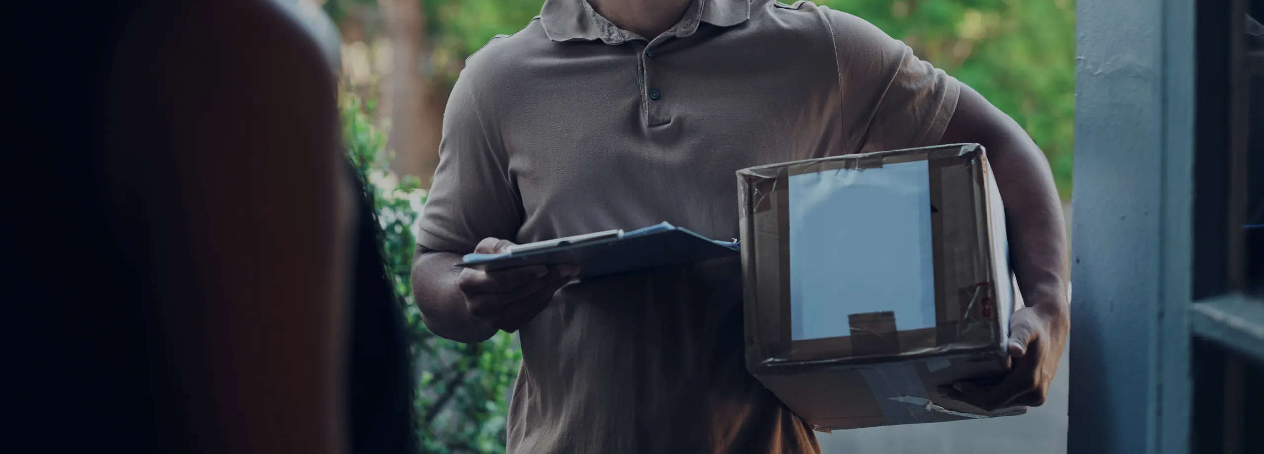 Um entregador amigável, com barba e boné, sorri enquanto entrega uma encomenda na porta de uma casa. Ele segura uma caixa de papelão em uma mão e uma prancheta na outra, interagindo com a pessoa que…