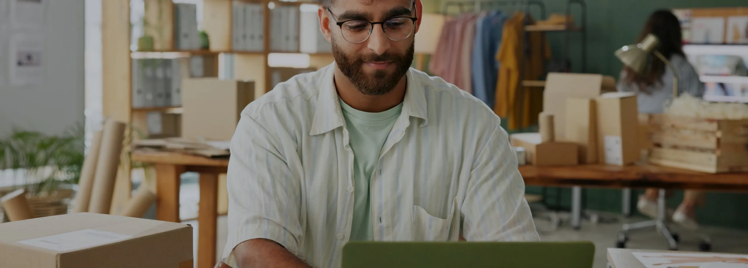 Um homem de barba e óculos trabalha concentrado e com um leve sorriso em seu laptop. Ele está no escritório de uma pequena empresa ou e-commerce, com caixas de embalagem, prateleiras e uma arara de r…