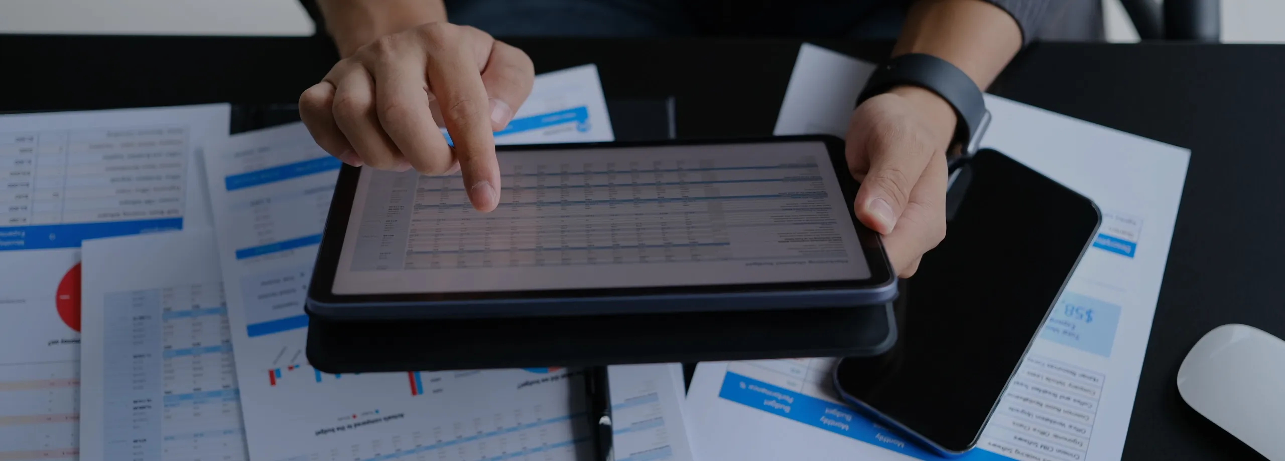 Um homem utilizando um tablet, está sentado em frente a uma mesa e nessa há várias folhas papeis com gráficos imprimidos nelas