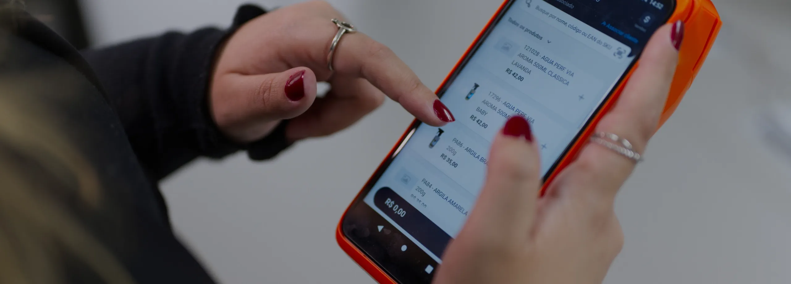Close-up das mãos de uma pessoa segurando e interagindo com um smartphone com capa laranja. A pessoa está usando o dedo indicador para navegar por um aplicativo de vendas ou estoque que exibe uma lis…