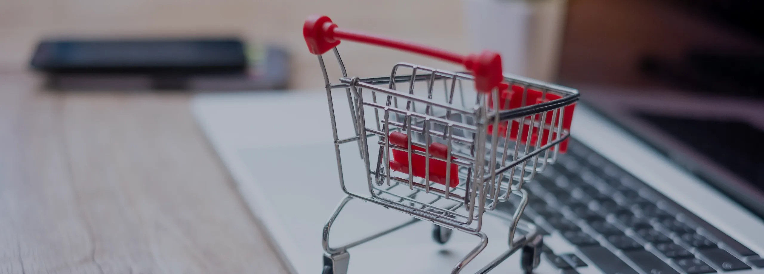Um close-up de um carrinho de compras em miniatura, feito de metal prateado e plástico rosa, que está parado em cima de um laptop aberto. O carrinho está ligeiramente inclinado para a direita e seus…