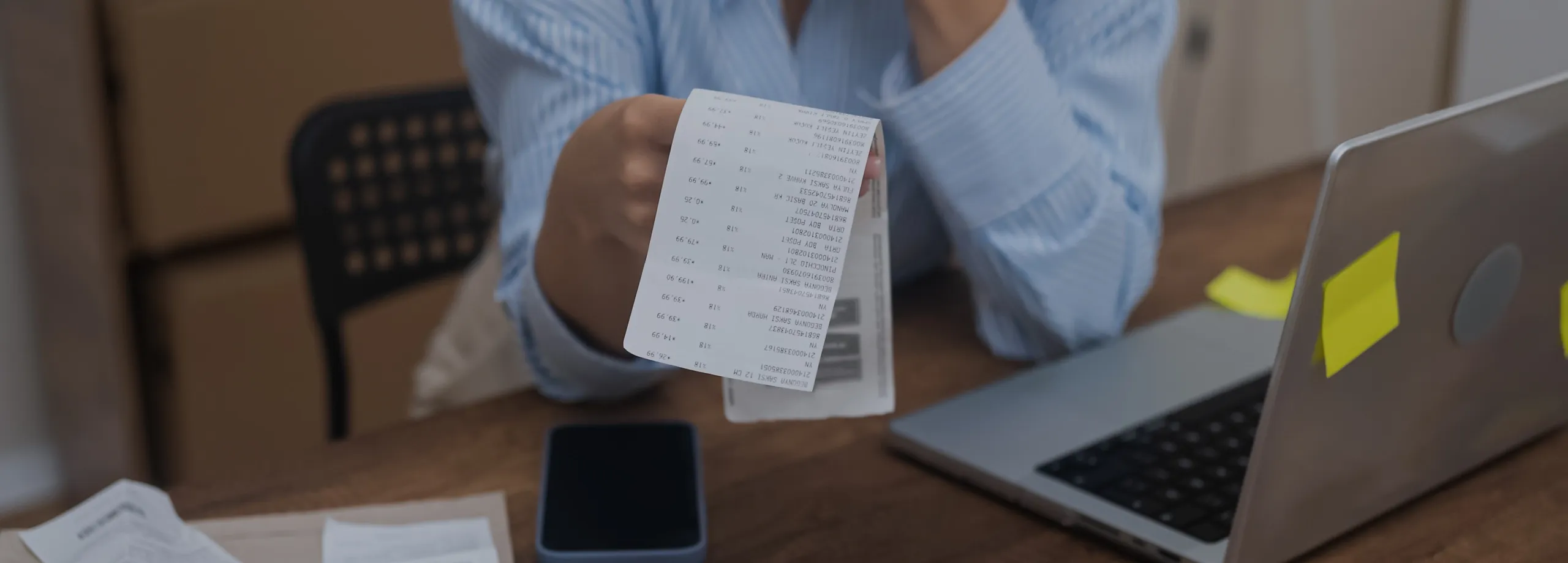 Uma pessoa sentada em uma mesa de madeira está segurando um recibo de papel branco com o braço estendido em direção ao observador. A pessoa está usando uma camisa listrada azul e branca. Na mesa, há…