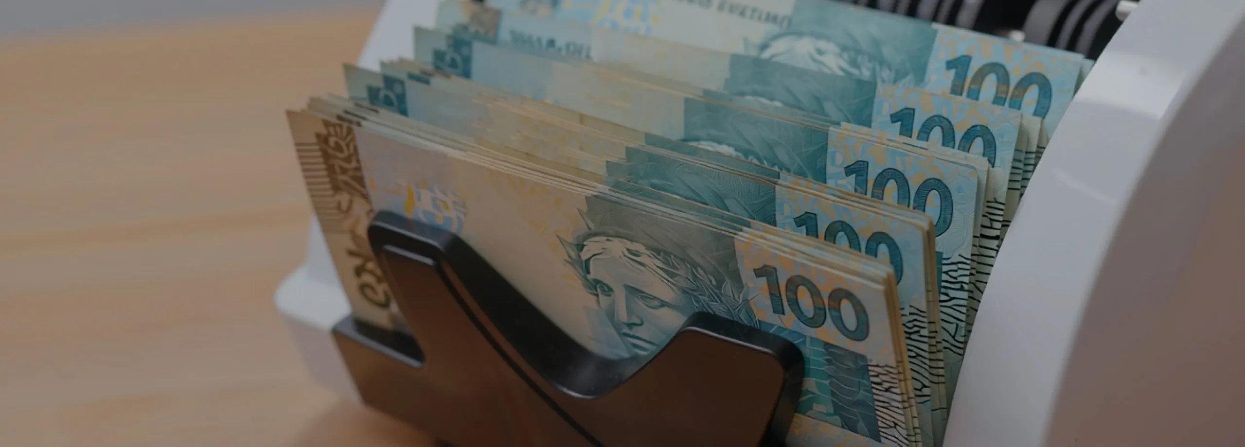 Um close-up de várias notas de 100 Reais sendo processadas por uma máquina de contar dinheiro. As notas, predominantemente azuis e beges, estão alinhadas e empilhadas na abertura da máquina, com a Ef…