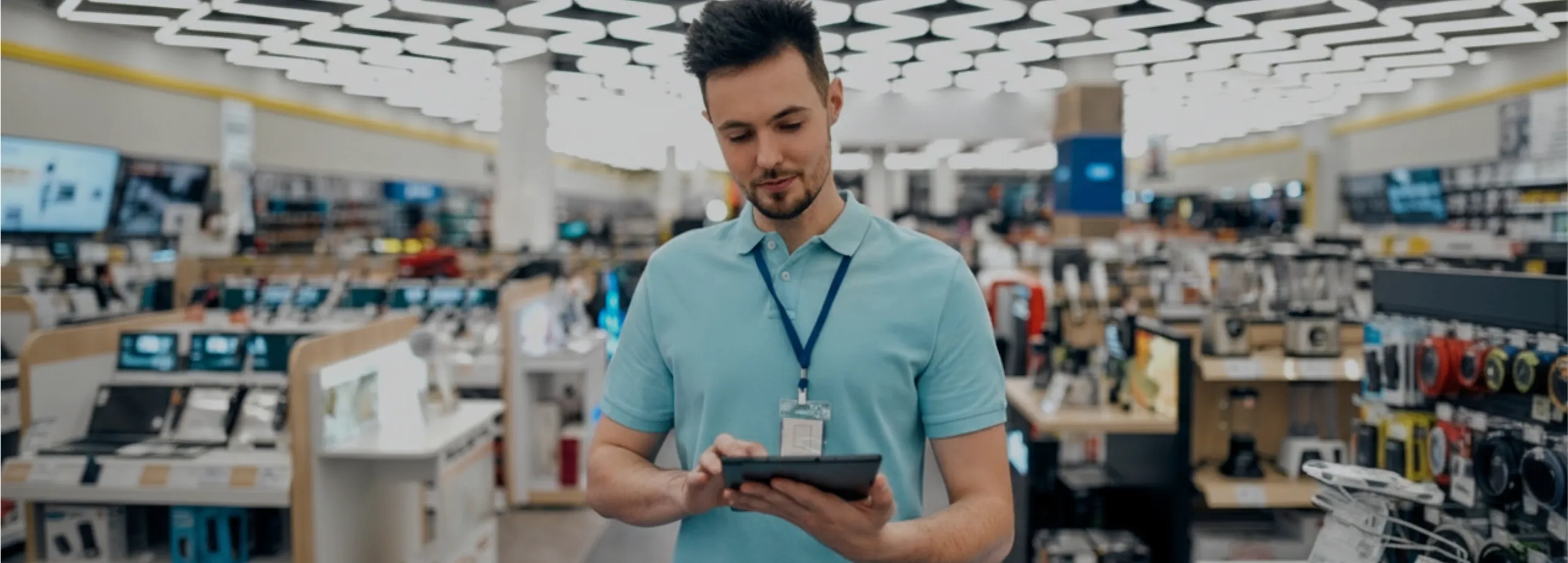 Um jovem vendedor em uma loja de eletrônicos, vestindo uma camisa polo azul clara e um crachá de identificação, está olhando para baixo e interagindo com um tablet digital que ele segura com as duas…