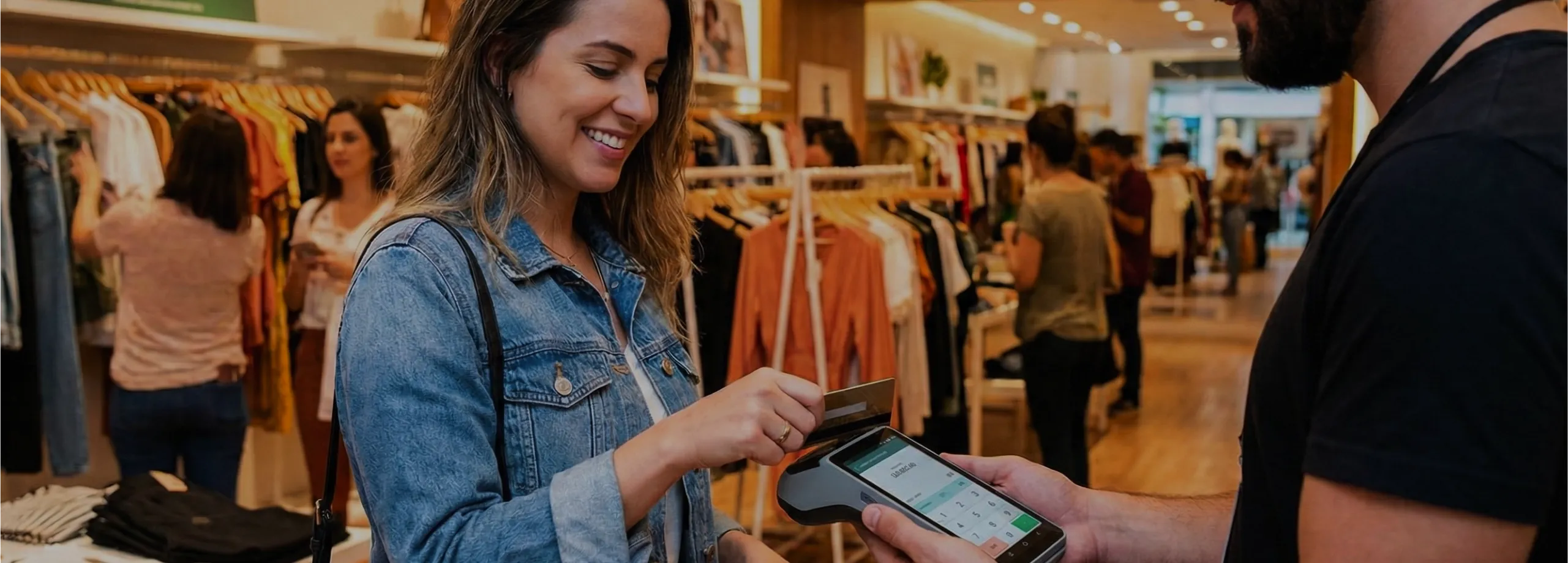 Em uma loja de roupas bem iluminada, uma mulher de cabelos longos e jaqueta jeans sorri enquanto insere um cartão de crédito em um terminal de pagamento móvel. Um vendedor de barba e camiseta preta,…