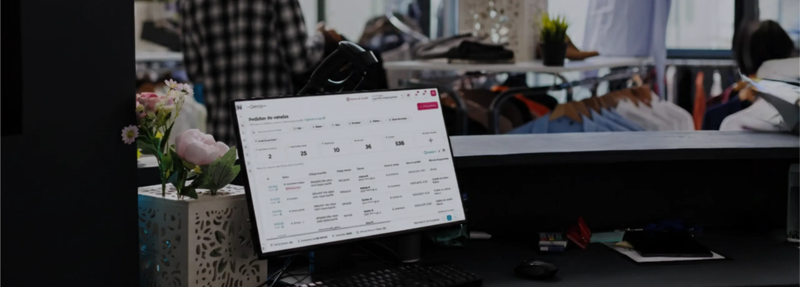 Em primeiro plano, sobre um balcão preto, um monitor exibe um sistema de gestão (dashboard) com números e listas de dados. Ao lado, há um teclado, mouse e um pequeno vaso decorativo branco com flores…
