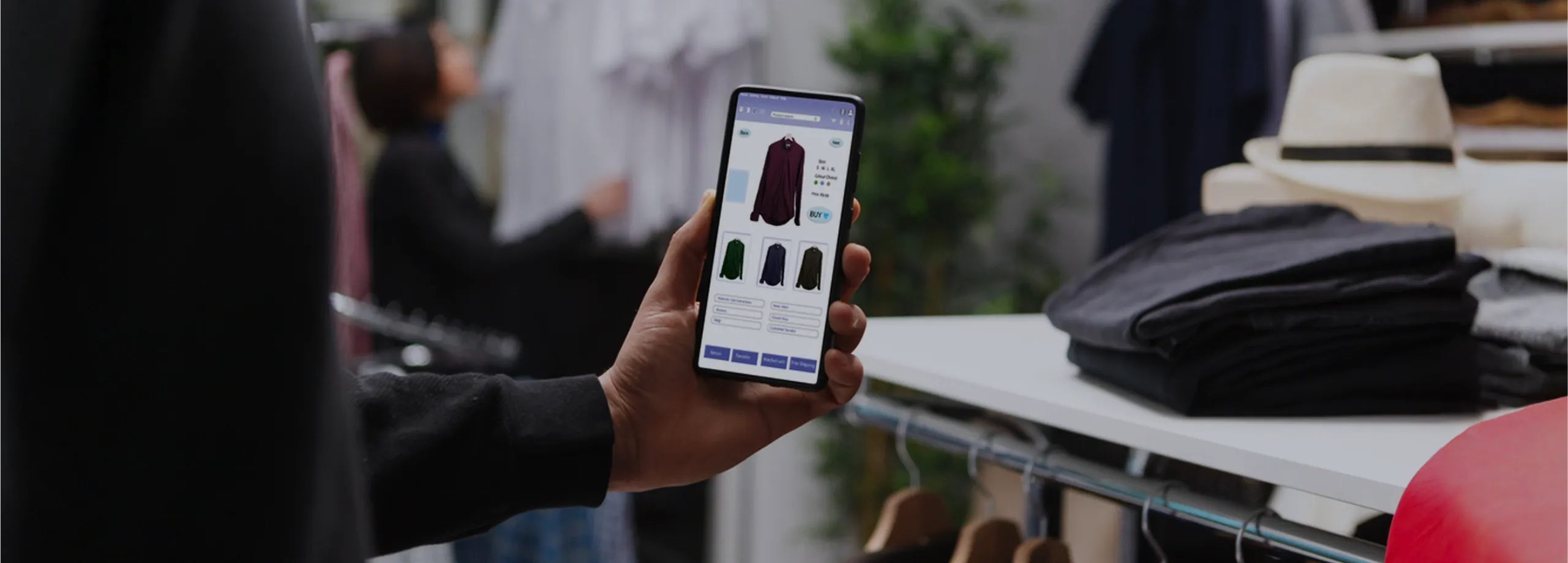 Em primeiro plano, a mão de uma pessoa segura um celular que exibe a interface de uma loja online, com a foto de uma camisa vinho e botão de compra. O cenário é o interior de uma loja física: à direi…