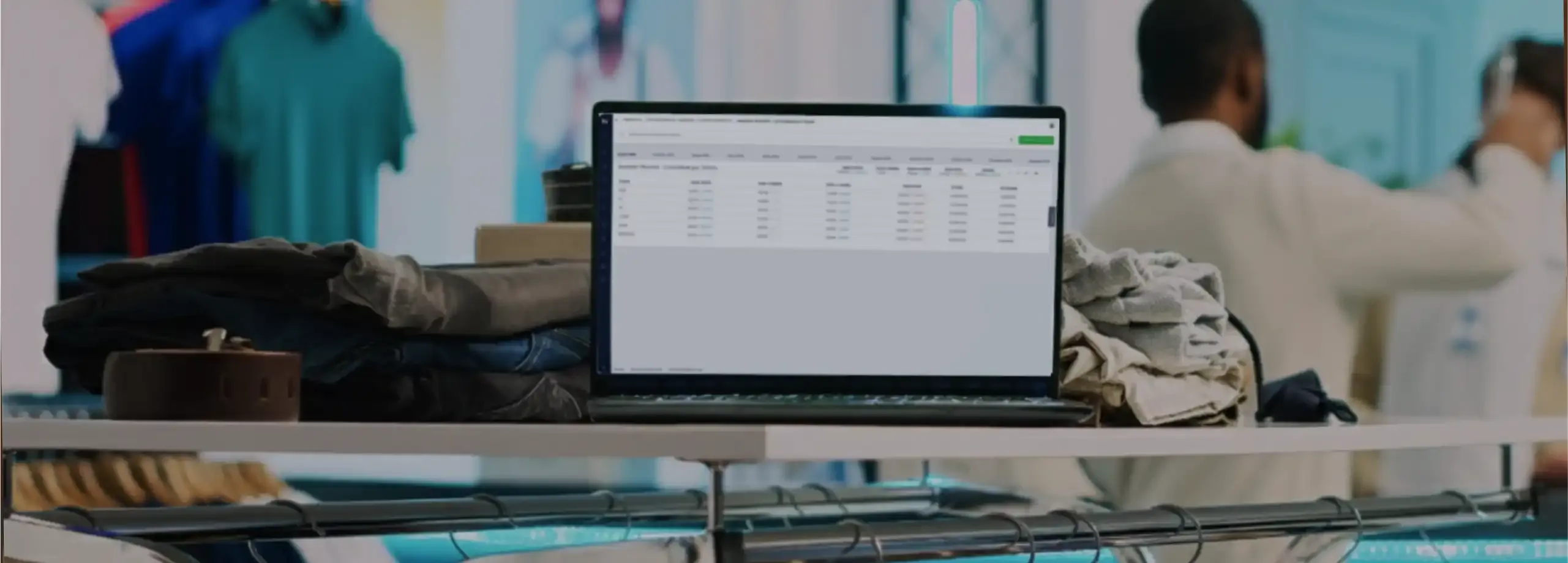 Em primeiro plano, um notebook está apoiado sobre uma prateleira branca, cercado por pilhas de calças jeans e roupas bege dobradas. A tela exibe uma tabela de dados de um software corporativo. Abaixo…