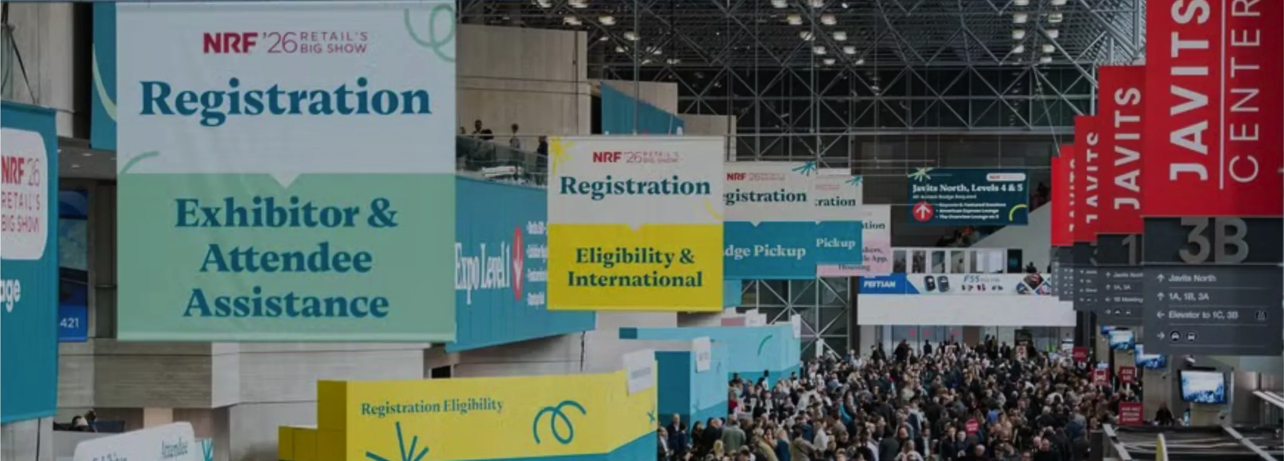 Uma visão ampla do interior do Javits Center repleto de milhares de participantes. No teto alto, pendem vários banners grandes de sinalização para o evento 'NRF '26 Retail's Big Show'. Um banner verd…