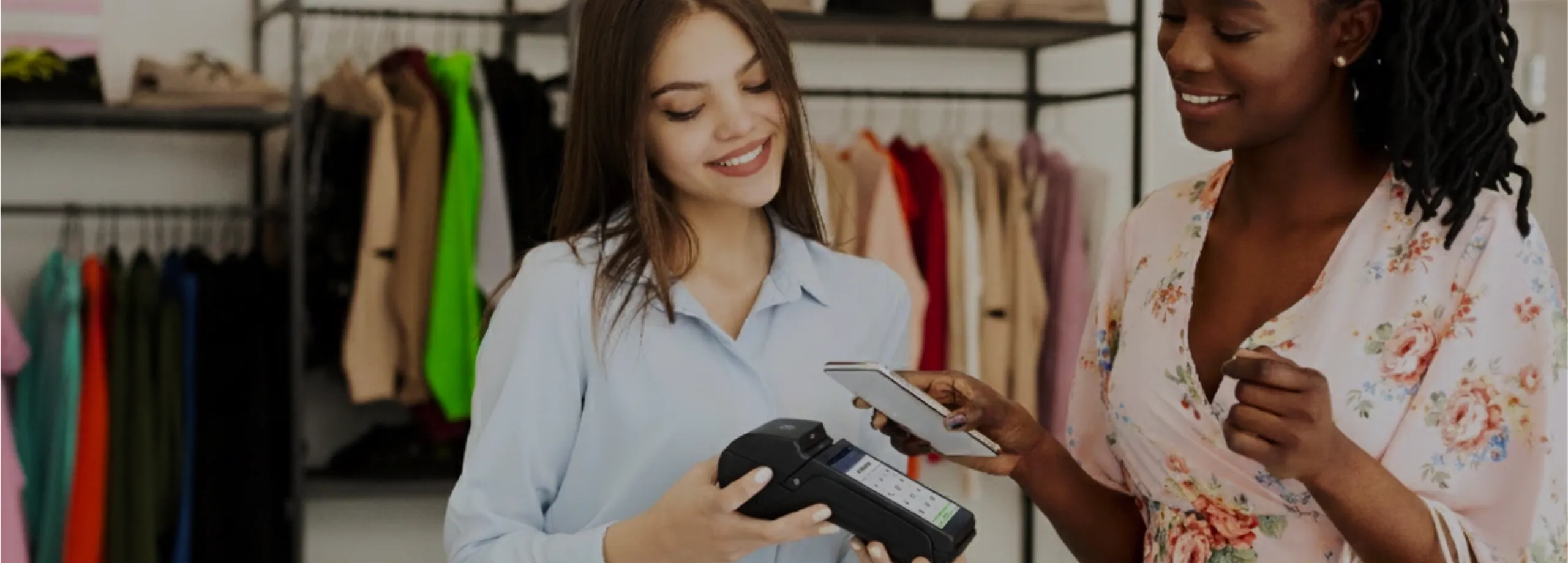 Em uma loja de roupas, uma vendedora de camisa azul clara (à esquerda) segura um terminal de pagamento móvel. À direita, uma cliente negra com tranças longas e blusa floral rosa sorri enquanto aproxi…