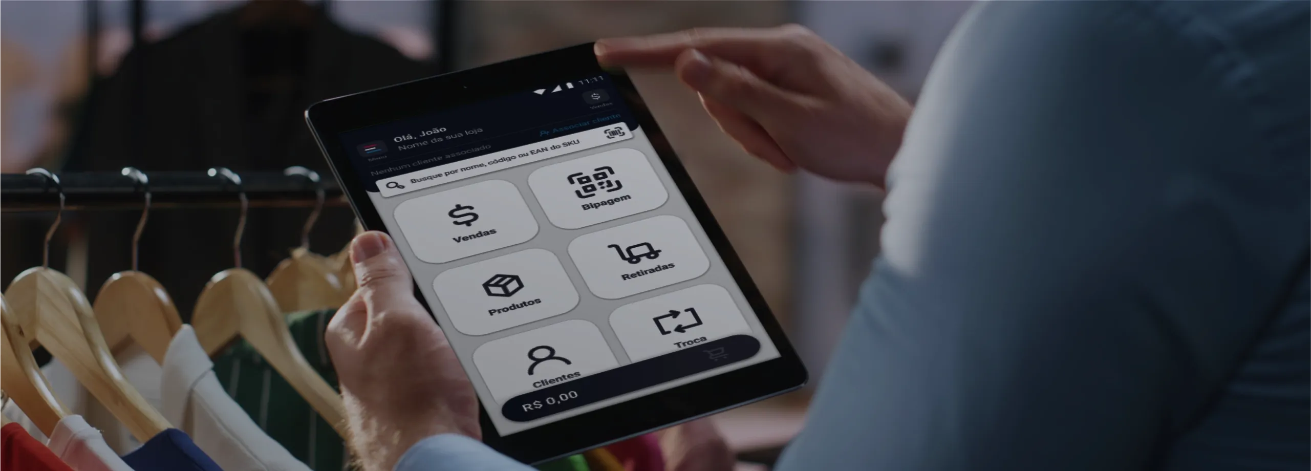 Close-up das mãos de um funcionário de camisa azul segurando um tablet. A tela exibe o painel de um aplicativo de varejo com botões grandes para: Vendas, Bipagem, Produtos, Retiradas, Clientes e Troc…