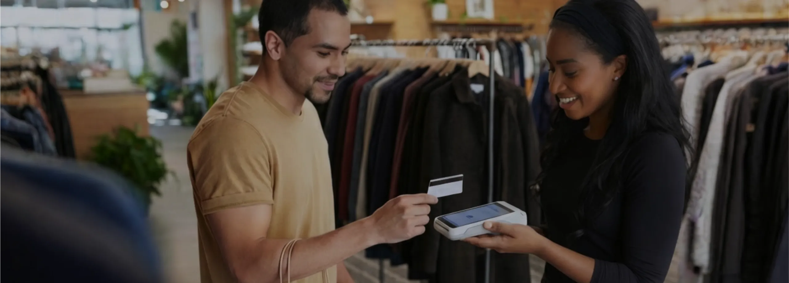 Cena em uma loja de roupas: à esquerda, um cliente de camiseta mostarda segura uma sacola de papel e estende um cartão de crédito. À direita, uma vendedora negra, vestida de preto e usando uma faixa…