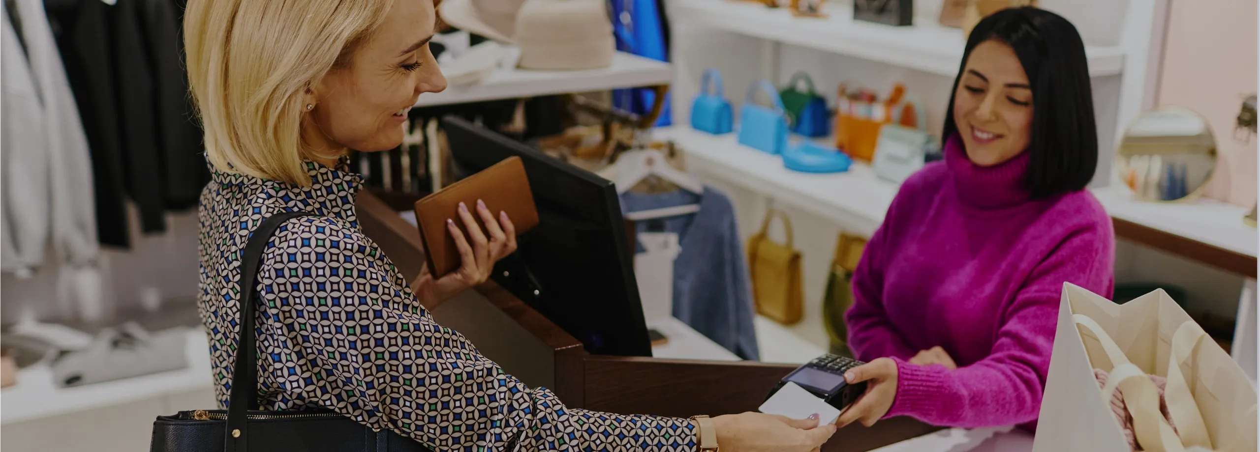 Cena no caixa de uma loja de moda. À esquerda, uma cliente loira de blusa estampada e bolsa preta no ombro sorri ao inserir um cartão branco no terminal de pagamento. Na outra mão, ela segura uma car…