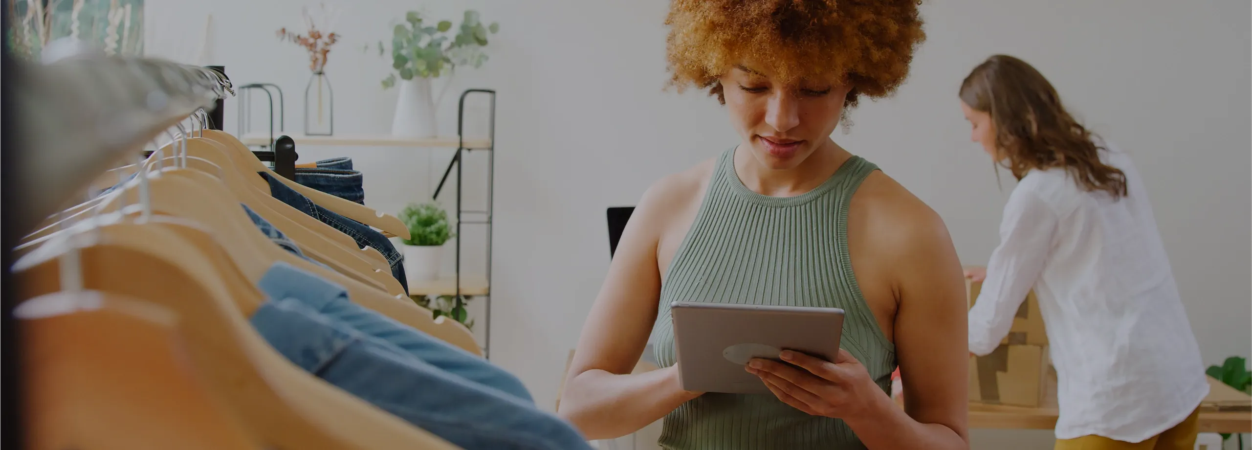 Uma mulher com cabelo afro volumoso usa um tablet em uma loja de roupas, com uma arara de cabides em primeiro plano e outra pessoa organizando caixas ao fundo.