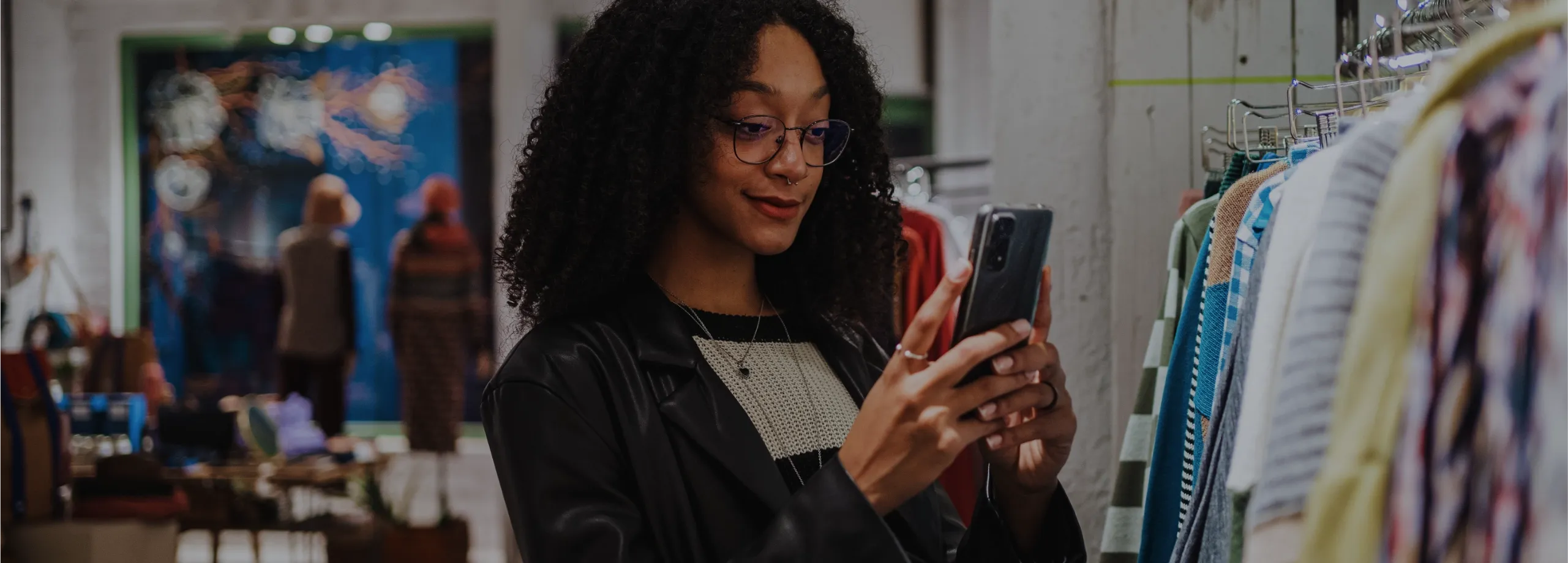 Mulher jovem com cabelos cacheados e óculos usa um smartphone em uma loja de roupas, próxima a uma arara de peças coloridas.
