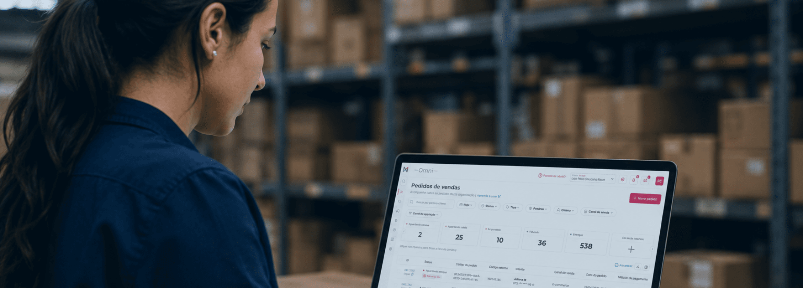 Mulher usando um notebook com um sistema de gestão de pedidos aberto, em um centro de distribuição com prateleiras cheias de caixas ao fundo.