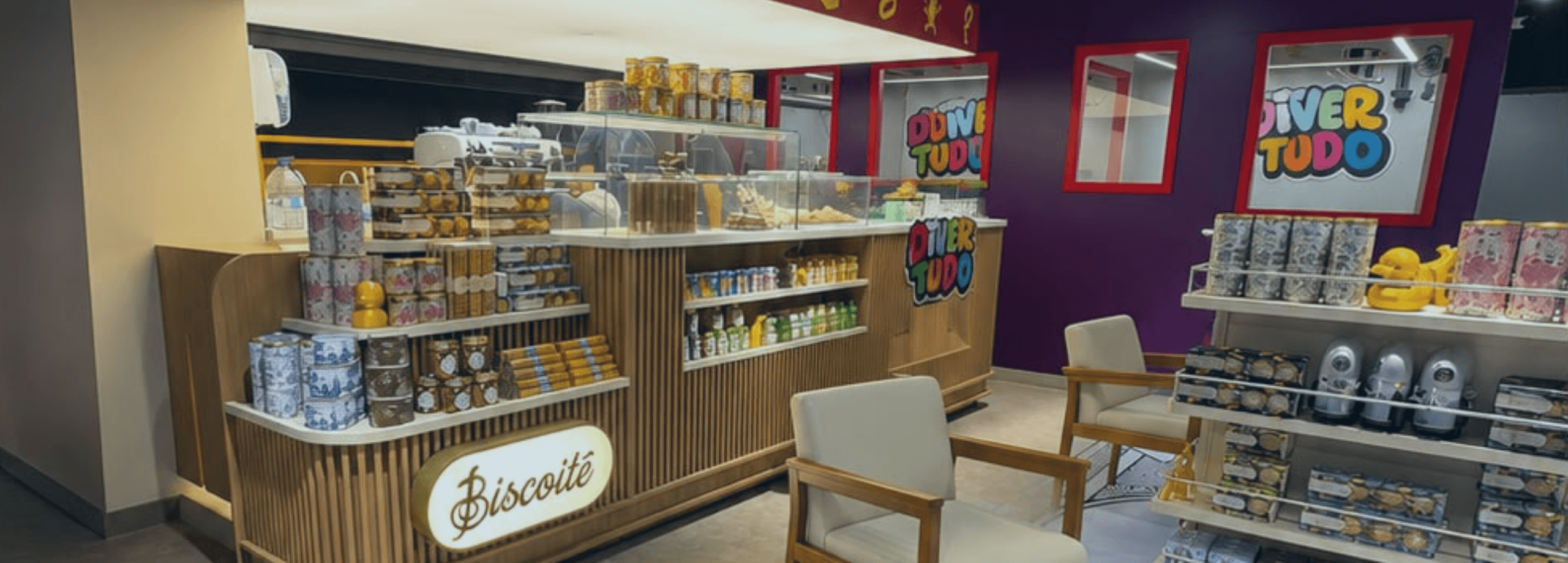 Fotografia do interior de uma loja conceito exibindo o balcão iluminado do café Biscoitê em madeira, poltronas claras e prateleiras com brinquedos e produtos sob a marca "Divertudo".
